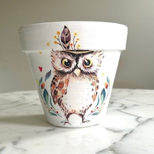 Puede incluir: Maceta de cerámica blanca decorada con una ilustración de búho en acuarela. El búho tiene plumas marrones y blancas, ojos grandes amarillos y un tocado de plumas. La maceta está adornada con detalles florales y de hojas en verde, rojo y amarillo.