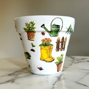 May include: White ceramic flower pot with a gardening theme. The pot is decorated with images of plants, a watering can, a fence, and yellow rain boots.