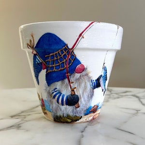 Może przedstawiać: Biała ceramiczna doniczka z kolorową ilustracją gnoma łowiącego ryby na plaży. Gnom ma na sobie niebieski kapelusz, niebiesko-białą w paski koszulę i trzyma wędkę.