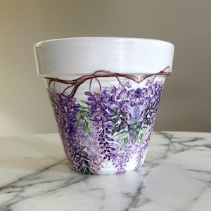 May include: White ceramic flower pot with a painted design of purple wisteria flowers and green leaves. The pot has a white rim and a decorative floral pattern around the body. A beautiful container for plants.