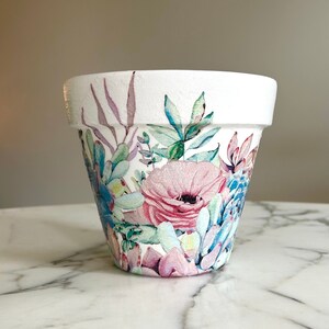 May include: White ceramic flower pot with a watercolour floral design featuring pink, blue, and green flowers and leaves.