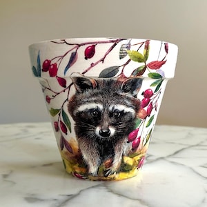 May include: A white ceramic flower pot with a painted design of a raccoon in a forest setting. The raccoon is looking directly at the viewer. The design includes branches, leaves, and red berries.