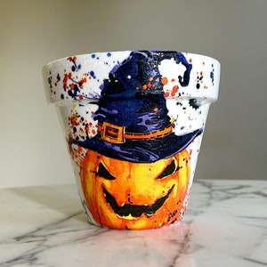 May include: A white ceramic planter with a hand-painted design of a pumpkin wearing a witch's hat. The pumpkin has a scary face and the hat is purple with a black brim. The planter is decorated with orange, yellow, and blue paint splatters.