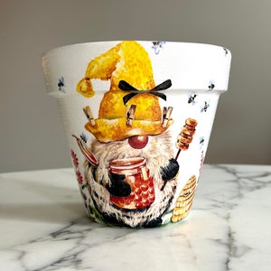 Puede incluir: Maceta de cerámica blanca con un diseño pintado a mano de un gnomo con un sombrero amarillo y sosteniendo un tarro de miel. El gnomo está rodeado de abejas y flores.
