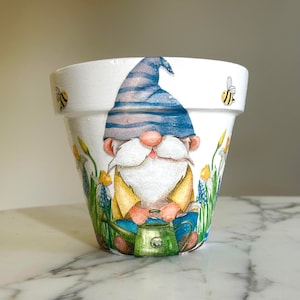 Pode incluir: Pote de flores de cerâmica branca com um desenho pintado à mão de um gnomo segurando um regador. O gnomo está usando um chapéu listrado azul e branco e uma camisa amarela. Há flores amarelas e azuis ao redor do gnomo e duas abelhas no pote.