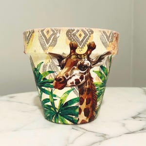 May include: A decorative flower pot featuring a giraffe design. The pot has a cream-colored background with a geometric pattern at the top and green tropical leaves at the bottom. The giraffe has brown spots and is surrounded by green leaves.