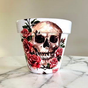 Puede incluir: Maceta de cerámica blanca con un diseño de calavera y rosas rojas.