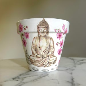 May include: White ceramic planter with a hand-painted design of a meditating Buddha surrounded by pink flowers and white stones.