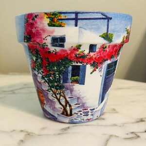 Puede incluir: Una maceta de cer&aacute;mica con una escena pintada de un edificio blanco con contraventanas y puertas azules, y flores rojas. La maceta tiene un borde azul y una superficie similar al m&aacute;rmol.