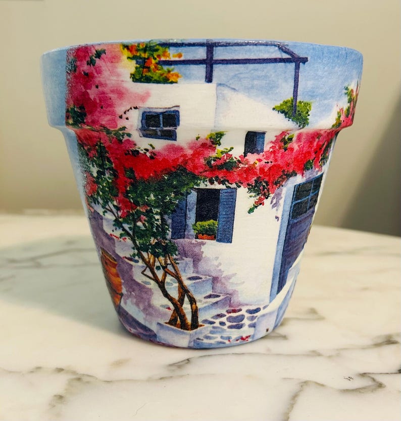 Puede incluir: Una maceta de cer&aacute;mica con una escena pintada de un edificio blanco con contraventanas y puertas azules, y flores rojas. La maceta tiene un borde azul y una superficie similar al m&aacute;rmol.