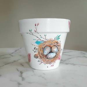 May include: White ceramic planter with a bird's nest design featuring three blue eggs. The design includes pink and green floral accents.