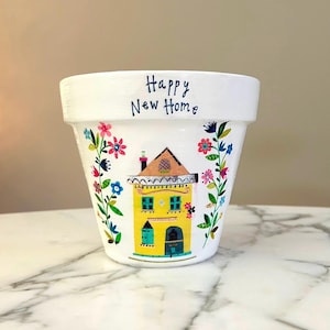 May include: White ceramic flower pot with a hand-painted yellow house design. The house has a brown roof and blue windows and door. The pot is decorated with colorful flowers and the words "Happy New Home" in blue script.