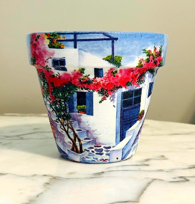 Puede incluir: Maceta decorativa con una escena pintada de un edificio blanco con detalles azules, una puerta azul y flores rojas. La maceta tiene una forma cl&aacute;sica y es adecuada para uso en interiores o exteriores. El dise&ntilde;o evoca una est&eacute;tica mediterr&aacute;nea.