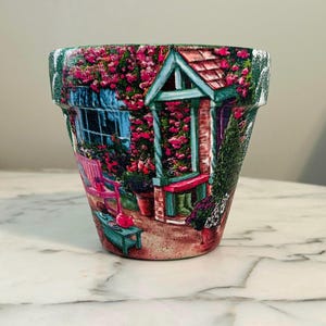 May include: A decorative terracotta flower pot featuring a detailed garden scene. The design includes a pink bench, a small table with a teapot, and a cottage with climbing roses. The pot is painted with vibrant colors and is suitable for indoor or outdoor use.