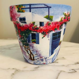 Puede incluir: Maceta de cer&aacute;mica pintada con una escena mediterr&aacute;nea. El dise&ntilde;o incluye un edificio blanco con contraventanas y puertas azules, una escalera de piedra y vibrantes flores rojas y rosas. La maceta es cil&iacute;ndrica.