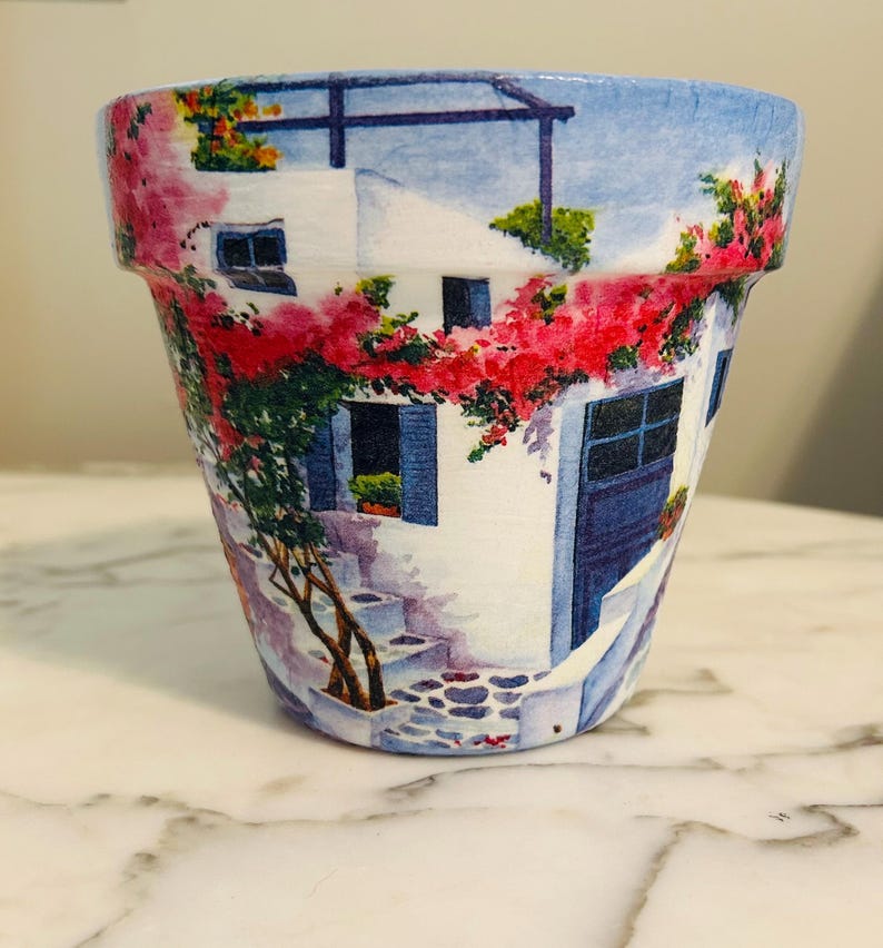 Puede incluir: Maceta de cer&aacute;mica pintada con una escena mediterr&aacute;nea. El dise&ntilde;o incluye un edificio blanco con contraventanas y puertas azules, una escalera de piedra y vibrantes flores rojas y rosas. La maceta es cil&iacute;ndrica.