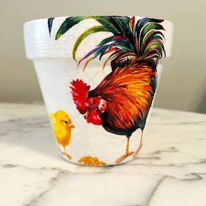 May include: White ceramic flower pot featuring a rooster and chick design. The rooster is red, orange, and yellow with a green, purple, and blue tail. The chick is yellow.