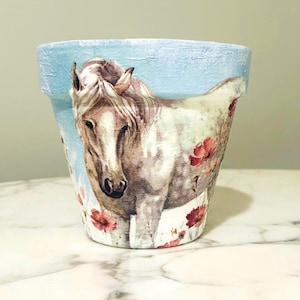 Könnte beinhalten: Hellblauer Keramik-Blumentopf mit einer Pferdeillustration und roten Blumen. Das Pferd ist weiß und braun, und die Blumen sind rot. Der Topf ist zylindrisch und für den Innen- oder Außenbereich geeignet.