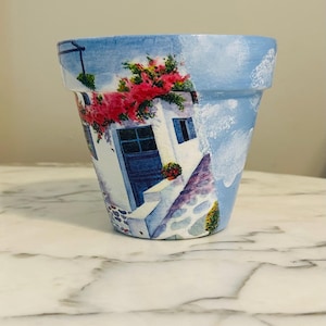 Puede incluir: Maceta de terracota pintada a mano con fondo de cielo azul. La maceta presenta un edificio blanco con puerta y ventanas azules, adornado con flores rojas. El dise&ntilde;o evoca una escena de pueblo mediterr&aacute;neo.