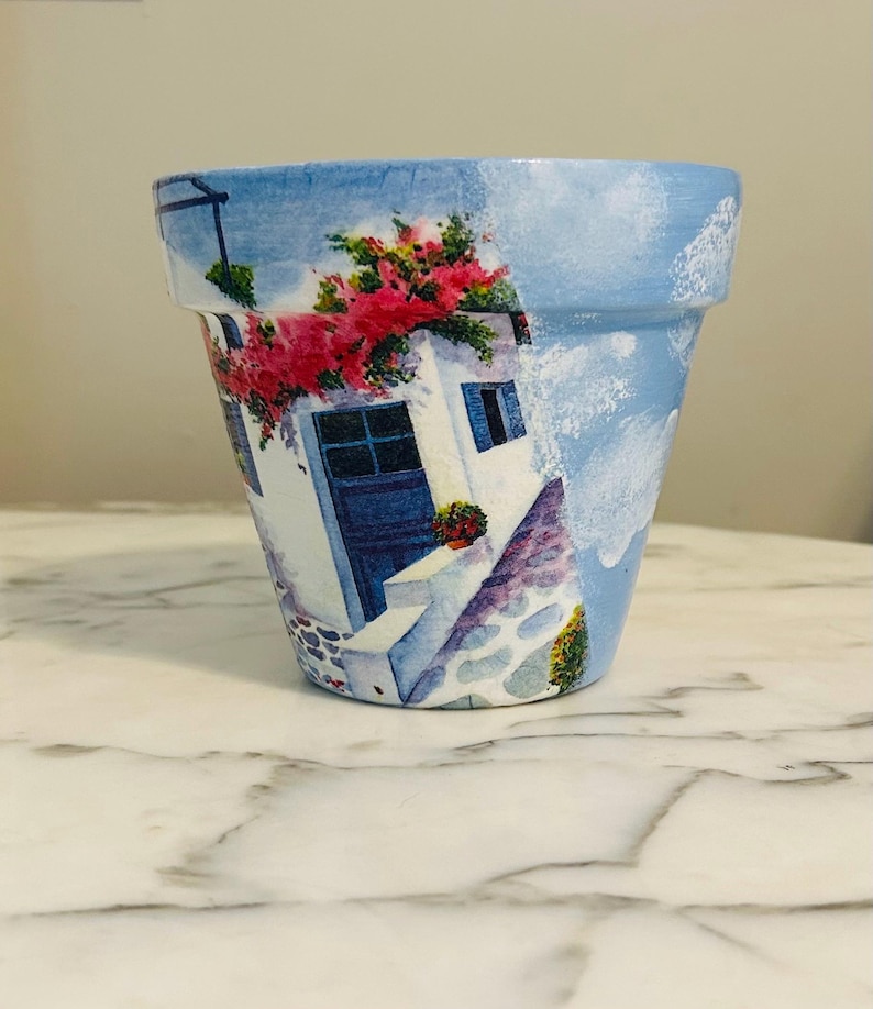 Puede incluir: Maceta de terracota pintada a mano con fondo de cielo azul. La maceta presenta un edificio blanco con puerta y ventanas azules, adornado con flores rojas. El dise&ntilde;o evoca una escena de pueblo mediterr&aacute;neo.