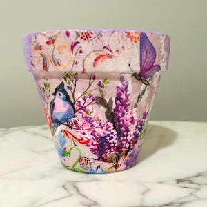 May include: A decorative flower pot with a floral and bird design. The pot features a lavender background with a bird, butterfly, and purple flowers. The rim of the pot is a lighter shade of lavender.