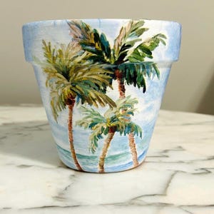 May include: A painted ceramic flower pot with a tropical beach scene. The pot is light blue with a design of palm trees, blue water, and a light blue sky. The palm trees have green fronds and brown trunks.