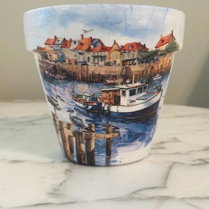 Może przedstawiać: Biała ceramiczna doniczka z kolorową ilustracją nadmorskiego miasteczka, łodzi i mew. Ilustracja jest namalowana w stylu akwarelowym.