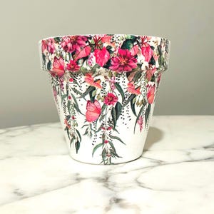 Op de afbeelding: Een witte keramische bloempot met een levendig bloemmotief. De pot heeft een herhalend patroon van roze bloemen, groene bladeren en vlinders. Het ontwerp loopt rond de hele pot, waardoor een decoratief en opvallend stuk voor planten ontstaat.