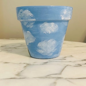 Puede incluir: Maceta de terracota pintada de azul claro con dise&ntilde;os de nubes blancas. La maceta es cil&iacute;ndrica con un borde ligeramente ensanchado. Las nubes tienen forma irregular y est&aacute;n dispersas por la superficie. La maceta est&aacute; vac&iacute;a.