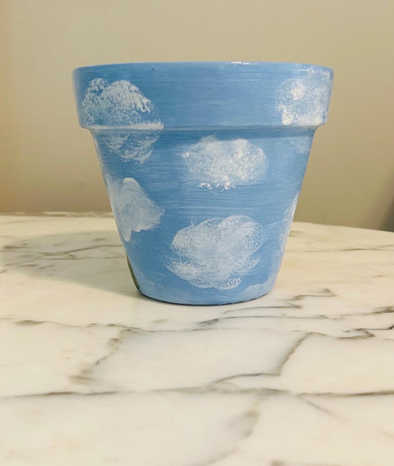 Puede incluir: Maceta de terracota pintada de azul claro con dise&ntilde;os de nubes blancas. La maceta es cil&iacute;ndrica con un borde ligeramente ensanchado. Las nubes tienen forma irregular y est&aacute;n dispersas por la superficie. La maceta est&aacute; vac&iacute;a.