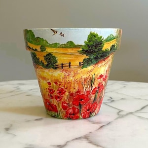 Peut inclure: Pot de fleur en terre cuite peint à la main, représentant un champ de coquelicots rouges. Le motif comprend un paysage jaune et vert avec une clôture, des arbres et des oiseaux sur fond de ciel bleu clair. Article décoratif pour la maison et le jardin.