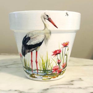 Może przedstawiać: Biała ceramiczna doniczka z szczegółową ilustracją bociana z czerwonymi nogami i dziobem, stojącego w pobliżu różowych lilii wodnych i zielonej trawy. Mała żaba jest widoczna w wodzie. Przedstawiono również ważkę.