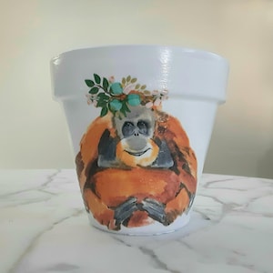 May include: White ceramic planter with a painted image of an orangutan wearing a floral crown. The orangutan is orange and brown with a gray face.