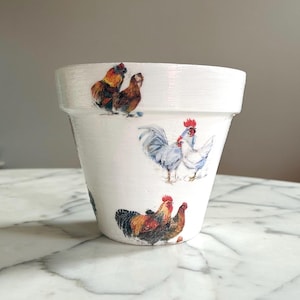 May include: White painted ceramic flower pot with rooster illustrations. The pot features multiple rooster designs in shades of red, brown, and blue. The pot is sitting on a marble surface.