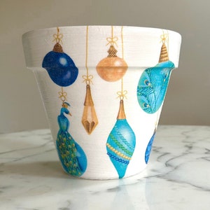 Puede incluir: Maceta de cerámica blanca con un diseño de adornos navideños azules, verdes y dorados. Los adornos cuelgan de cuerdas doradas con lazos.