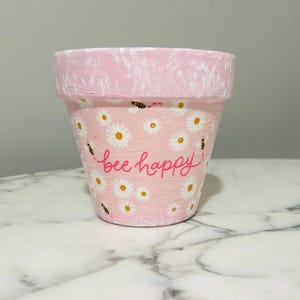 Puede incluir: Una maceta pintada de color rosa con un borde texturizado. La maceta está decorada con margaritas blancas, pequeñas abejas negras y la frase "bee happy" en escritura rosa. La maceta está diseñada para plantar flores.