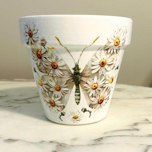 May include: White ceramic plant pot featuring a butterfly design. The butterfly wings are composed of daisies with yellow centres. The butterfly's body is green and brown. The pot sits on a white marble surface.