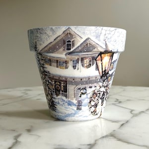 May include: A white ceramic planter with a winter scene design. The planter features a house with snow on the roof, a street lamp, and trees. The scene is painted in shades of blue, white, and brown.