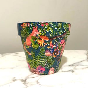 May include: A decorative teal-colored flower pot featuring a woodland design. The design includes a fox, birds, mushrooms, and foliage. The pot is cylindrical with a slightly flared rim, perfect for indoor or outdoor plants.