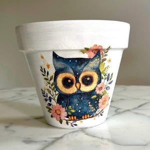 Puede incluir: Maceta de cerámica blanca con un búho azul pintado con estrellas y flores.