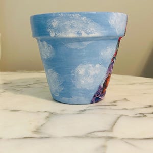 Puede incluir: Una maceta de terracota pintada a mano. La maceta est&aacute; pintada de azul claro con detalles de nubes blancas. Un dise&ntilde;o floral colorido es visible en el lateral de la maceta. La maceta est&aacute; sobre una superficie de m&aacute;rmol blanco.