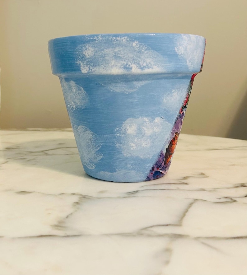 Puede incluir: Una maceta de terracota pintada a mano. La maceta est&aacute; pintada de azul claro con detalles de nubes blancas. Un dise&ntilde;o floral colorido es visible en el lateral de la maceta. La maceta est&aacute; sobre una superficie de m&aacute;rmol blanco.