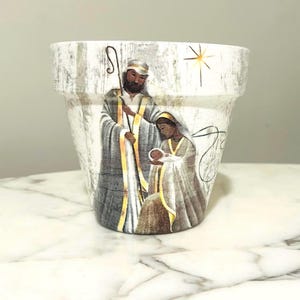 Könnte beinhalten: Weißer Keramik-Blumentopf mit einer gemalten Weihnachtskrippe. Das Bild zeigt Josef, Maria und das Jesuskind, mit einem Stern im Hintergrund. Die Figuren sind in Gewändern mit goldenen Akzenten gekleidet.