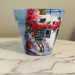 Puede incluir: Maceta decorativa de cer&aacute;mica con una escena pintada de un pueblo mediterr&aacute;neo. La maceta presenta un borde azul y una ilustraci&oacute;n detallada de un edificio blanco con flores rojas, contraventanas azules y un camino empedrado.