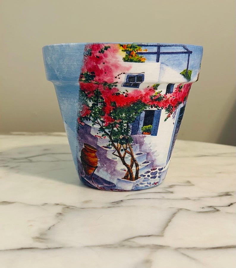 Puede incluir: Maceta decorativa de cer&aacute;mica con una escena pintada de un pueblo mediterr&aacute;neo. La maceta presenta un borde azul y una ilustraci&oacute;n detallada de un edificio blanco con flores rojas, contraventanas azules y un camino empedrado.