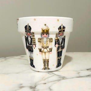 Puede incluir: Maceta de cerámica blanca decorada con tres figuras de cascanueces en negro, dorado y blanco. La maceta tiene un aspecto ligeramente envejecido con detalles de estrellas doradas.