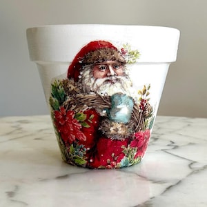 May include: White ceramic planter with a Christmas design featuring Santa Claus holding a mug, surrounded by red poinsettias and greenery.