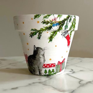 Könnte beinhalten: Weißer Keramiktopf mit einem weihnachtlichen Design mit einer Katze, einer Maus, einem Weihnachtsbaum und Geschenken. Das Design ist in Rot-, Grün- und Goldtönen gehalten.