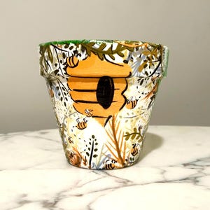 May include: A hand-painted terracotta flower pot featuring a beehive design. The pot is decorated with bees, foliage, and a honeycomb pattern in shades of orange, brown, green, and white. The rim of the pot is painted green.