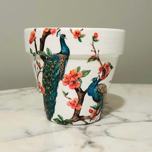 Könnte beinhalten: Weißer Keramik-Blumentopf mit einer detaillierten Malerei von zwei Pfauen auf blühenden Zweigen. Die Pfauen haben blaues und grünes Gefieder, und die Blüten sind leuchtend orange-rot. Der Topf ist ca. 15 cm hoch und für den Innen- und Außenbereich geeignet.